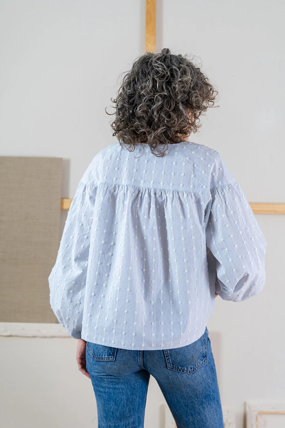 Rear view of a person wearing a light blue polka dot Positano blouse and jeans, standing in a room with neutral walls and a wooden frame.