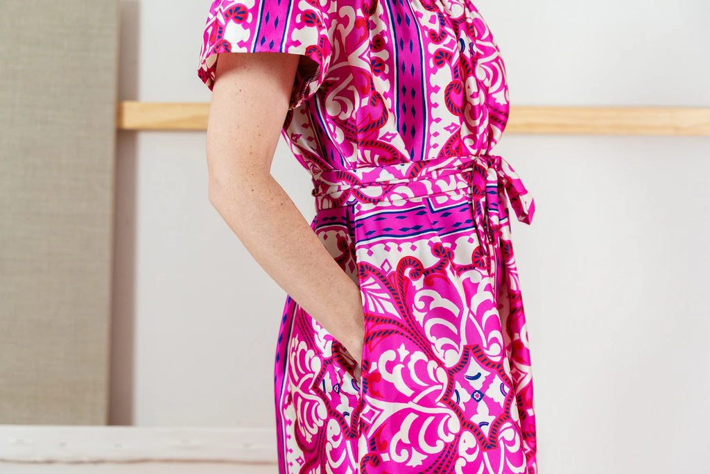 Close up of the waist of a person wearing a pink and purple patterned Positano dress standing in a minimal room.