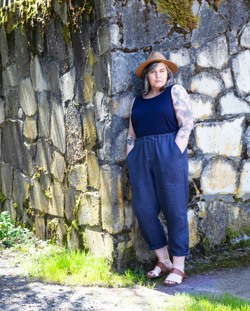 Person wearing navy Free Range Slacks, a navy tank top, and brown hat standing against a stone wall.