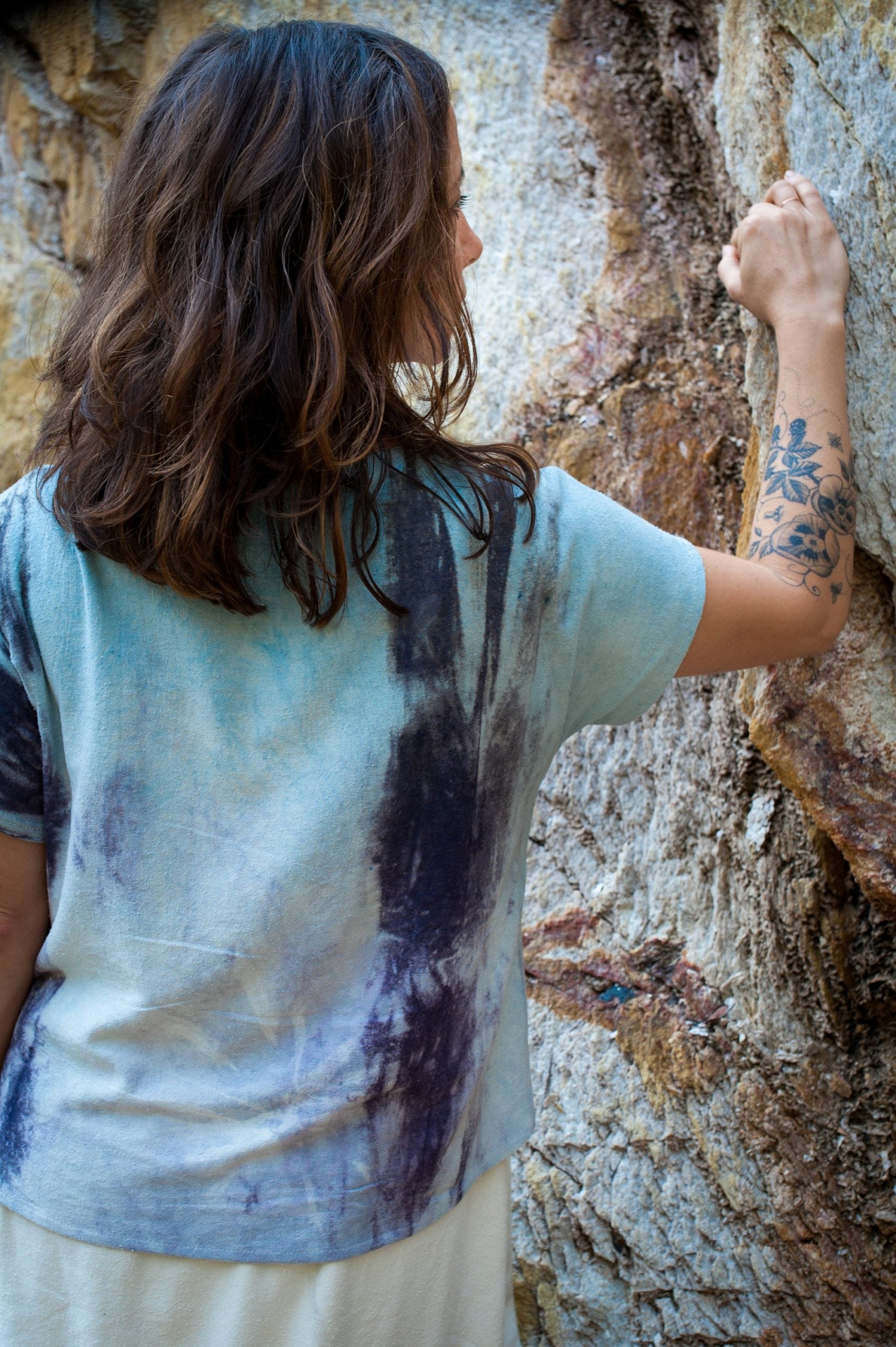 Person wearing a tie-dye shirt, seen from behind standing by a rock wall.