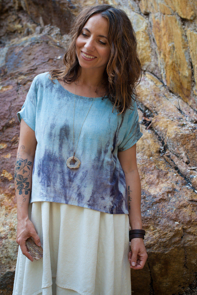 Woman wearing a tie-dye shirt and white skirt standing against a rocky wall.