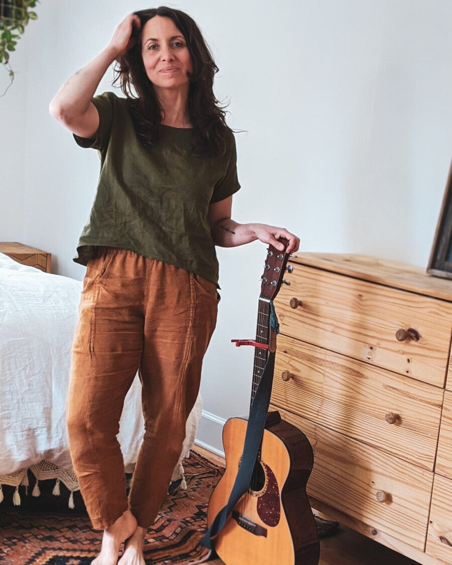 Woman standing in a bedroom with a guitar leaning against a dresser, wearing a short sleeve top.