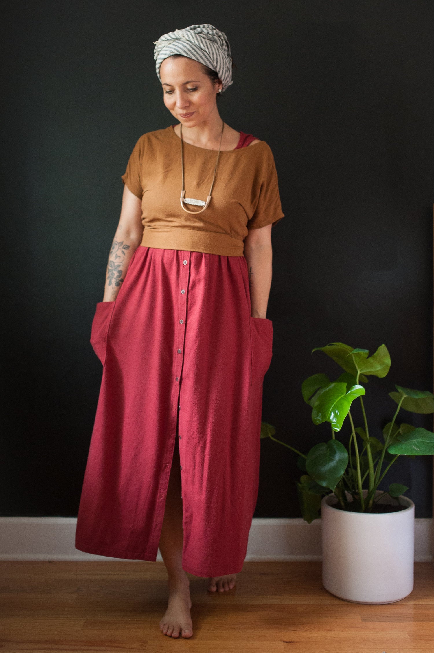 Woman wearing a short sleeve knit mustard top and red skirt standing in a room with a plant.