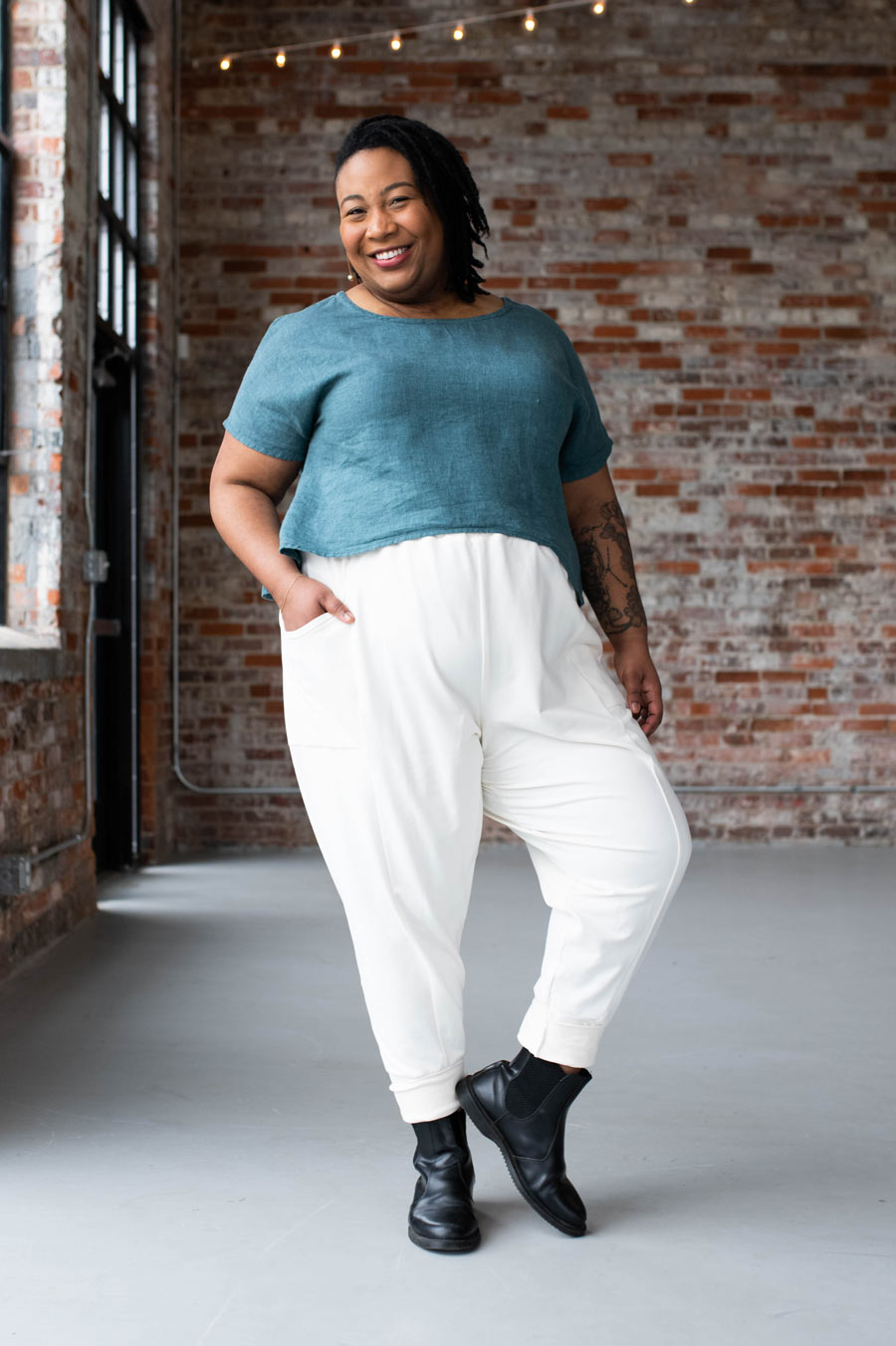 Person wearing a teal top and white pants standing in front of a brick wall.