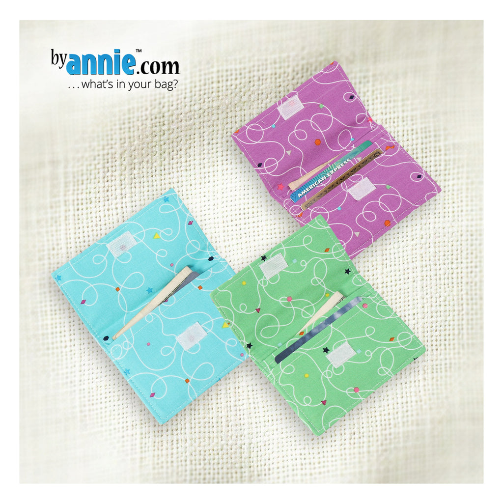 Three colorful fabric wallets with patterns on a textured surface, featuring 'byannie.com' branding.